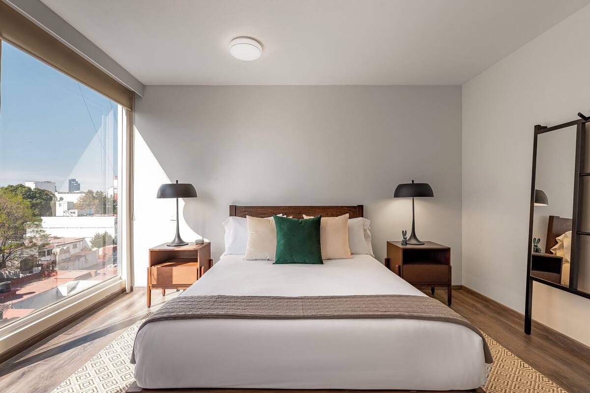 A modern hotel room with a large bed, two bedside tables with lamps, a large window with a city view, and a mirror on the wall.