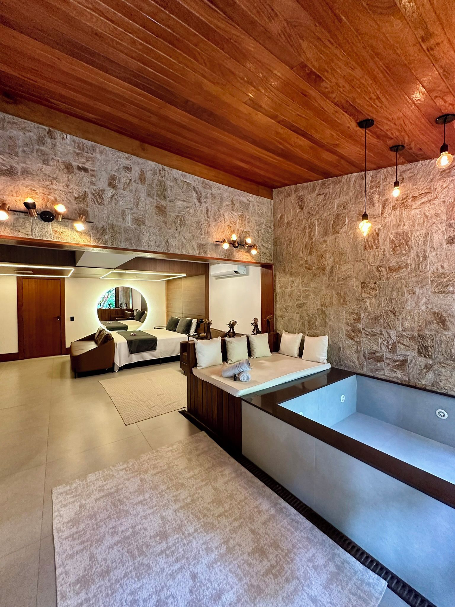 Un moderno spa con una bañera grande, techo de madera, paredes de piedra y una zona de estar con espejo.