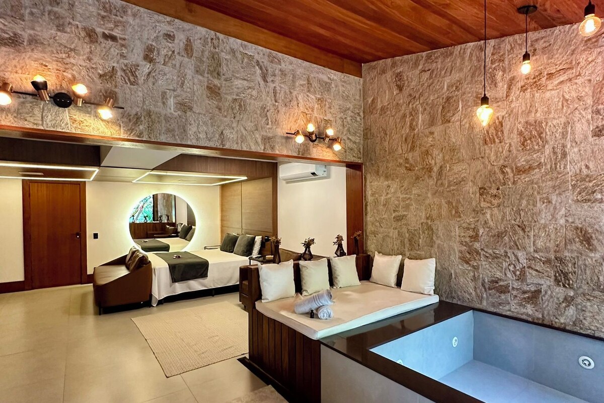 A modern spa room with a large bathtub, wooden ceiling, stone walls, and a seating area with a mirror.