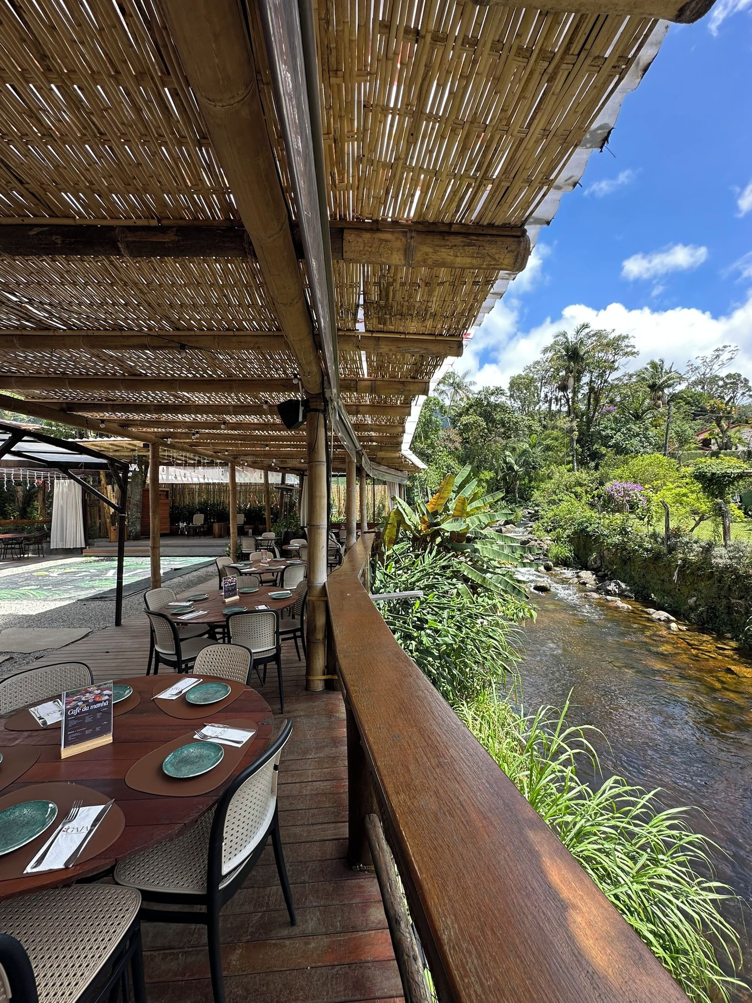 Ein Restaurant im Freien mit Tischen und Stühlen, einer Bambusdecke und Blick auf einen Bach mit viel Grün.