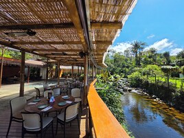 Un área de comedor al aire libre con mesas y sillas, techo de bambú y vistas a un río y follaje.
