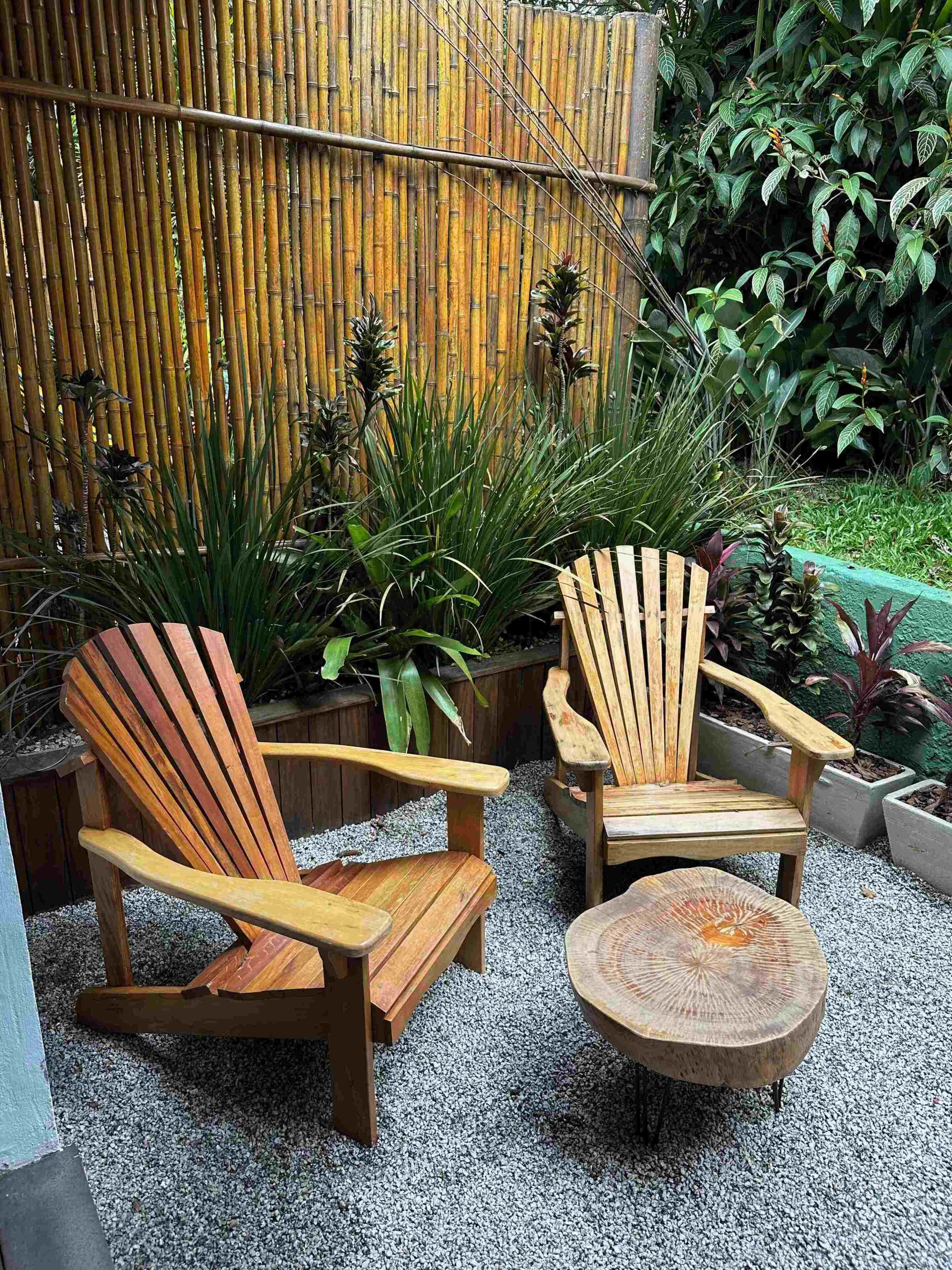 Two wooden Adirondack chairs and a round wooden table in a garden with a bamboo fence and various plants.