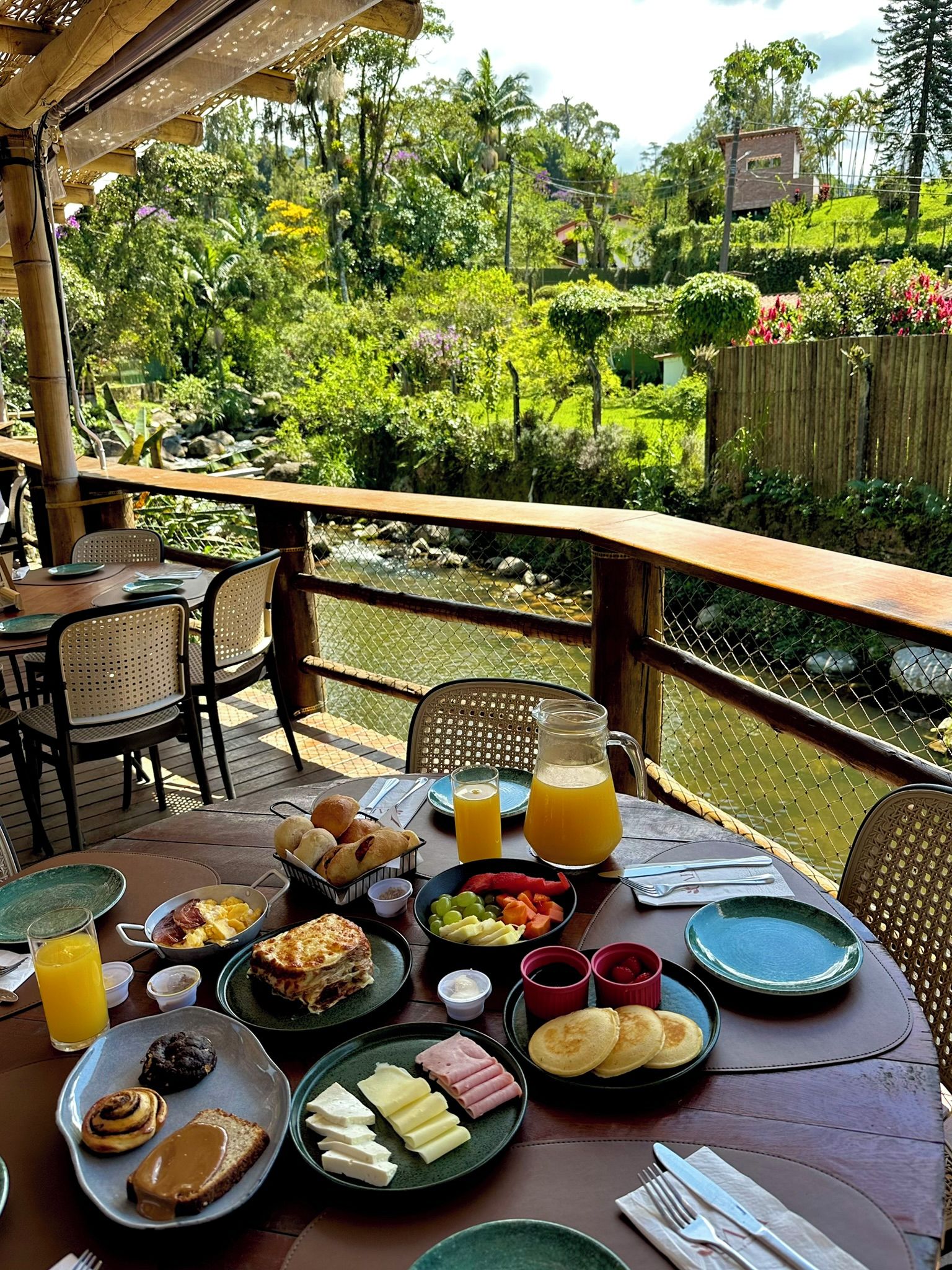 Un área de comedor al aire libre con una mesa lista para el desayuno, rodeada de abundante follaje y vistas a un jardín.