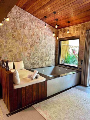 A modern bathroom with a stone wall, wooden ceiling, and a large bathtub. - Gaia Hotelaria e Gastronomia (Teresópolis)