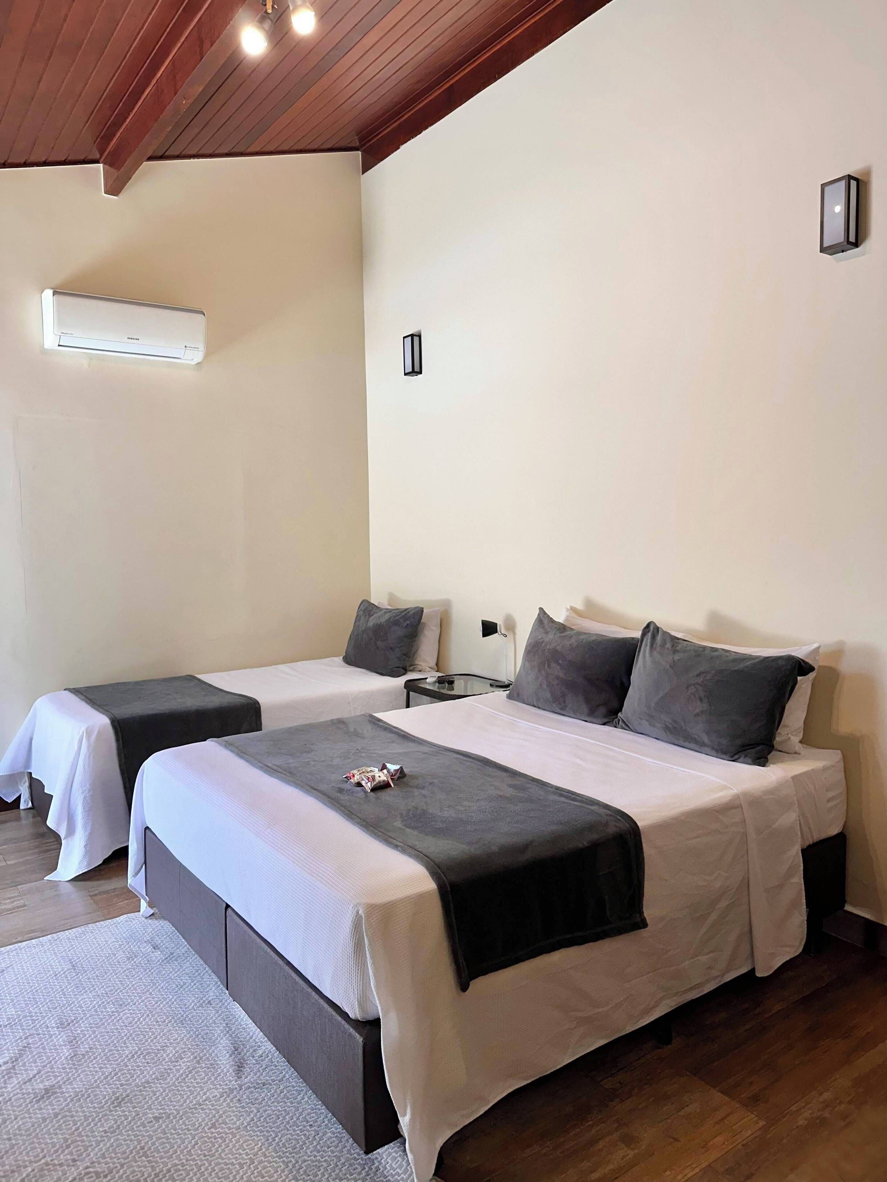 A hotel room with two beds, a ceiling fan, a wall-mounted air conditioner, and a bedside table with a lamp.