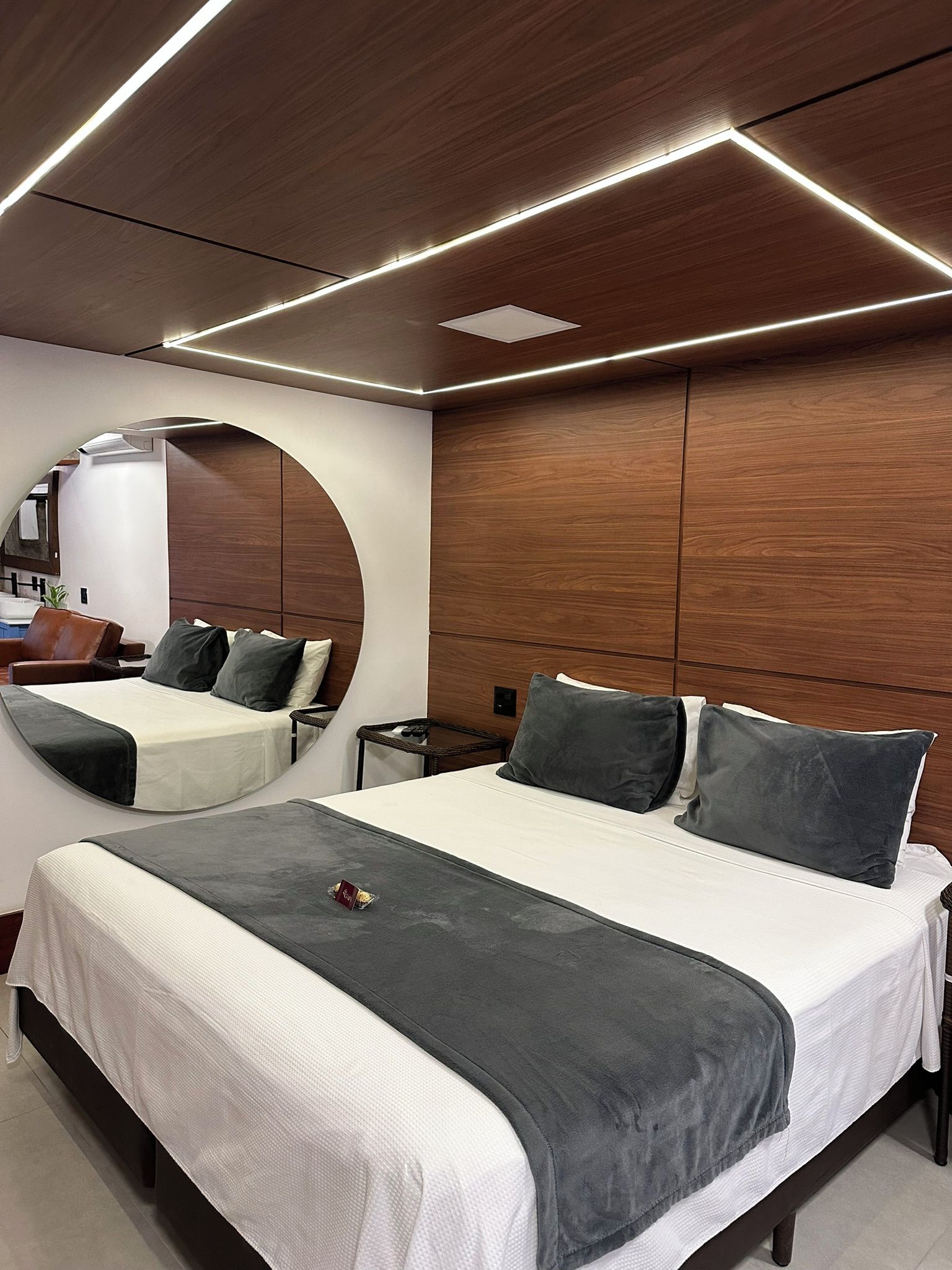A modern hotel room with a large bed, a round mirror, and a wooden panel wall.