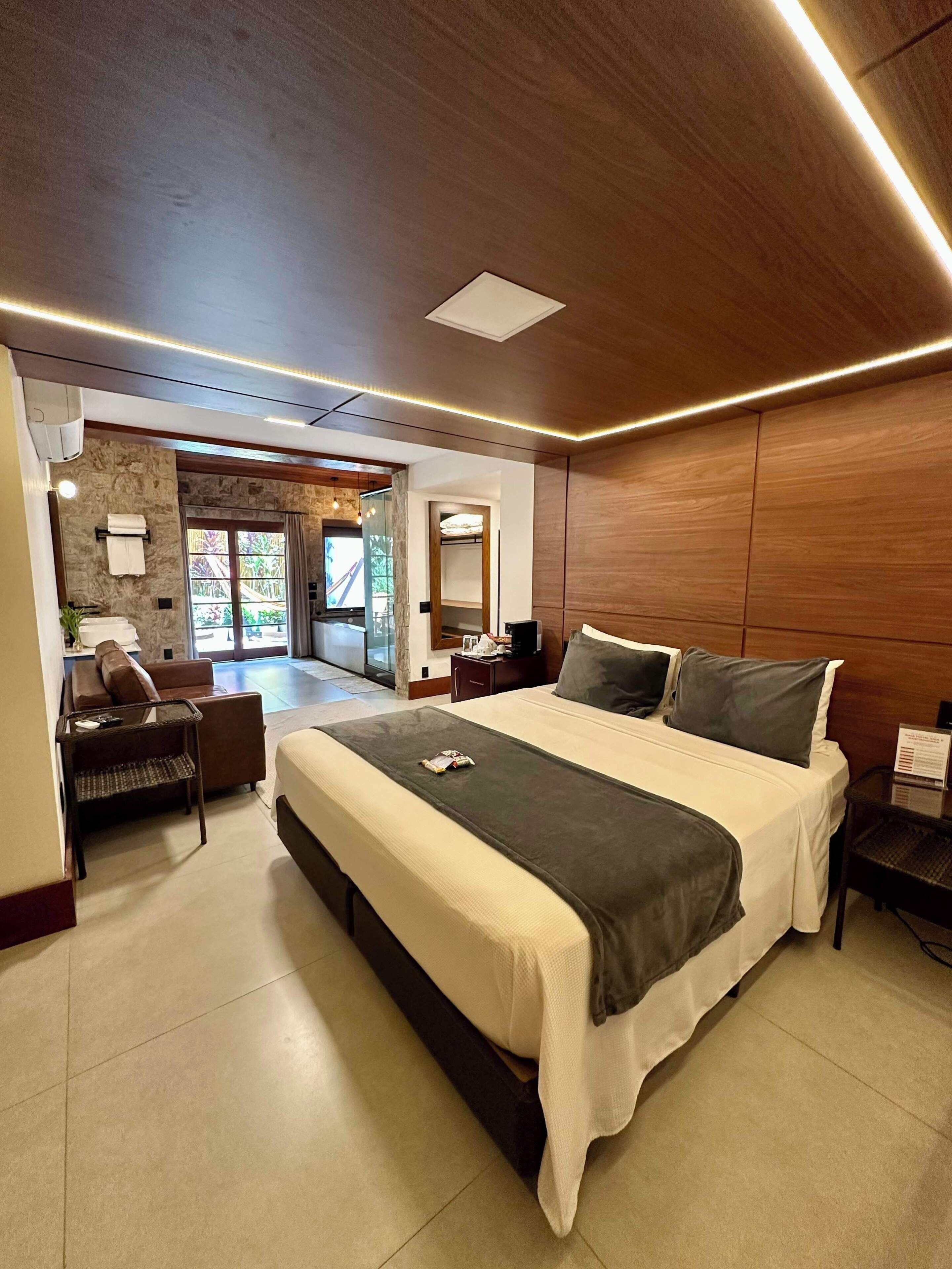 Una habitación de hotel moderna con una cama grande, una zona de estar con sofá y sillones, una mesita y vistas al exterior.