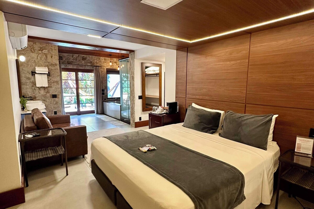 A modern hotel room with a large bed, a seating area with a sofa and armchairs, a small table, and a view of the outside.