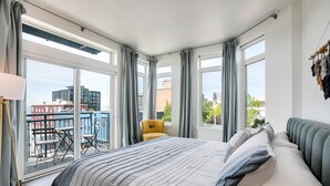 Habitación de hotel con una cama grande, cortinas grises, un balcón con una silla amarilla y vistas a edificios.