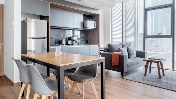 A modern kitchen with a built-in microwave, a stainless steel refrigerator, and a wooden dining table with chairs. There is a gray sofa with a throw blanket and a small wooden side table. - Blueground Josefa by Urbanistas CDMX (Mexico City)