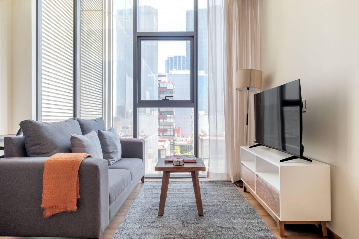 Ein modernes Wohnzimmer mit einem grauen Sofa, einem kleinen Holztisch, einem Fernseher auf einem weißen Standfuß und einem großen Fenster mit Blick auf die Stadt.
