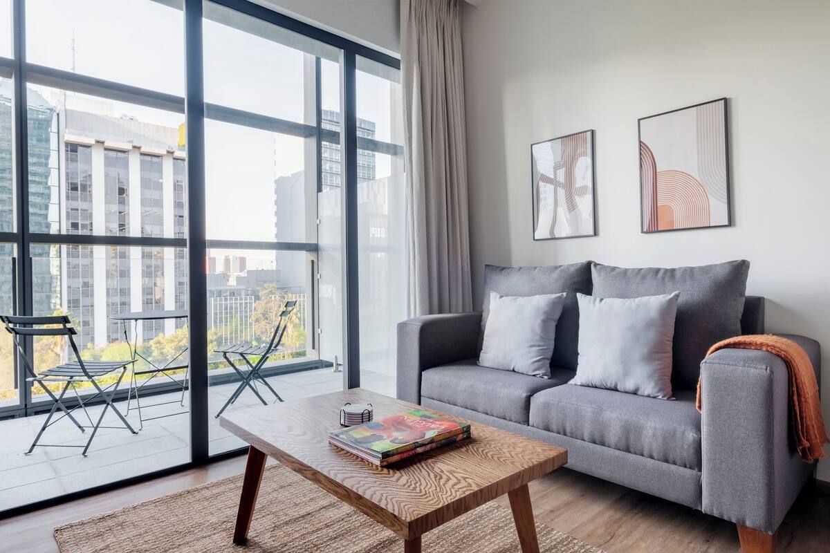 Ein modernes Wohnzimmer mit grauem Sofa, einem Holztisch und Blick auf Gebäude durch große Fenster.