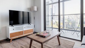 A modern living room with a flat-screen TV on a white and wooden cabinet, a coffee table with a magazine, and a large window with a city view. - Blueground Josefa by Urbanistas CDMX (Mexico City)
