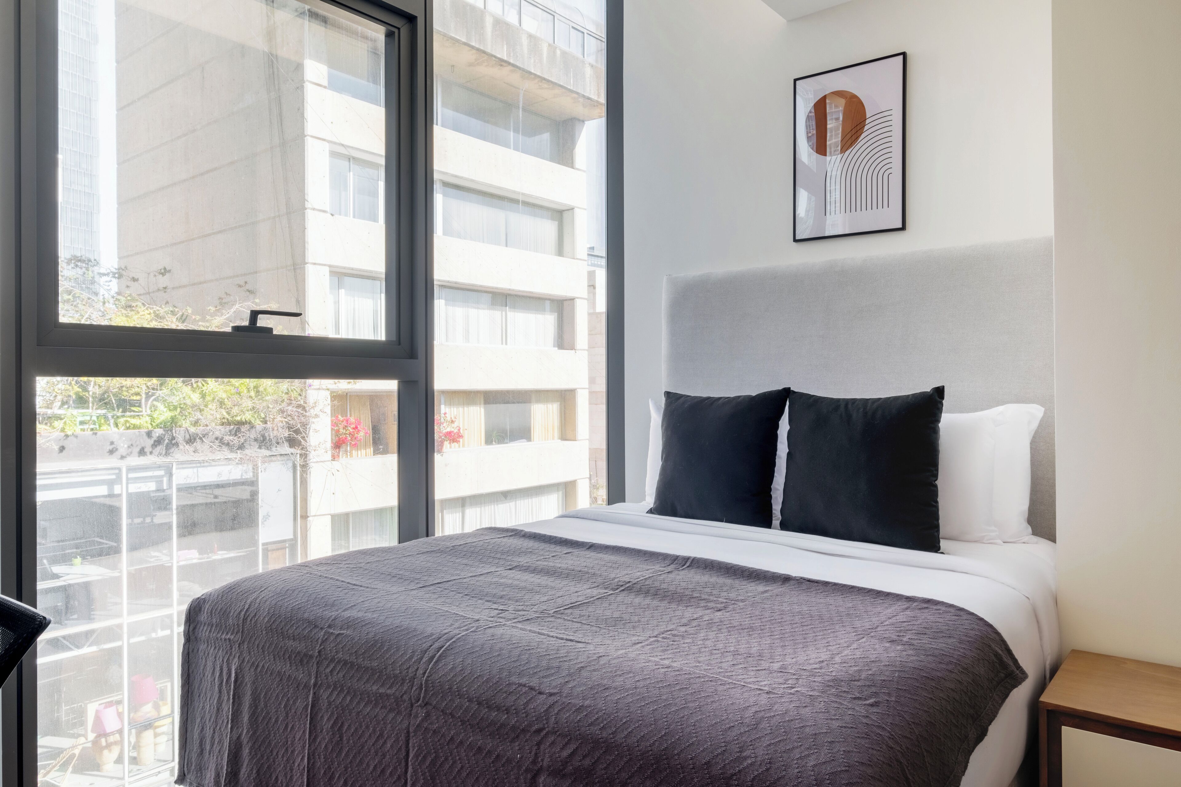 Ett hotellrum med ett stort fönster, en säng med ett grått överkast, två svarta kuddar och en inramad tavla på väggen.