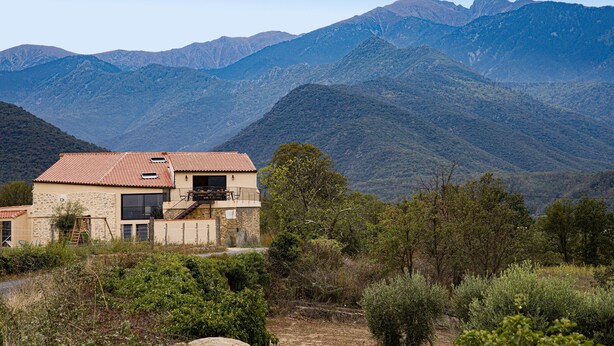A two-story house with a red-tiled roof, surrounded by mountains and greenery.