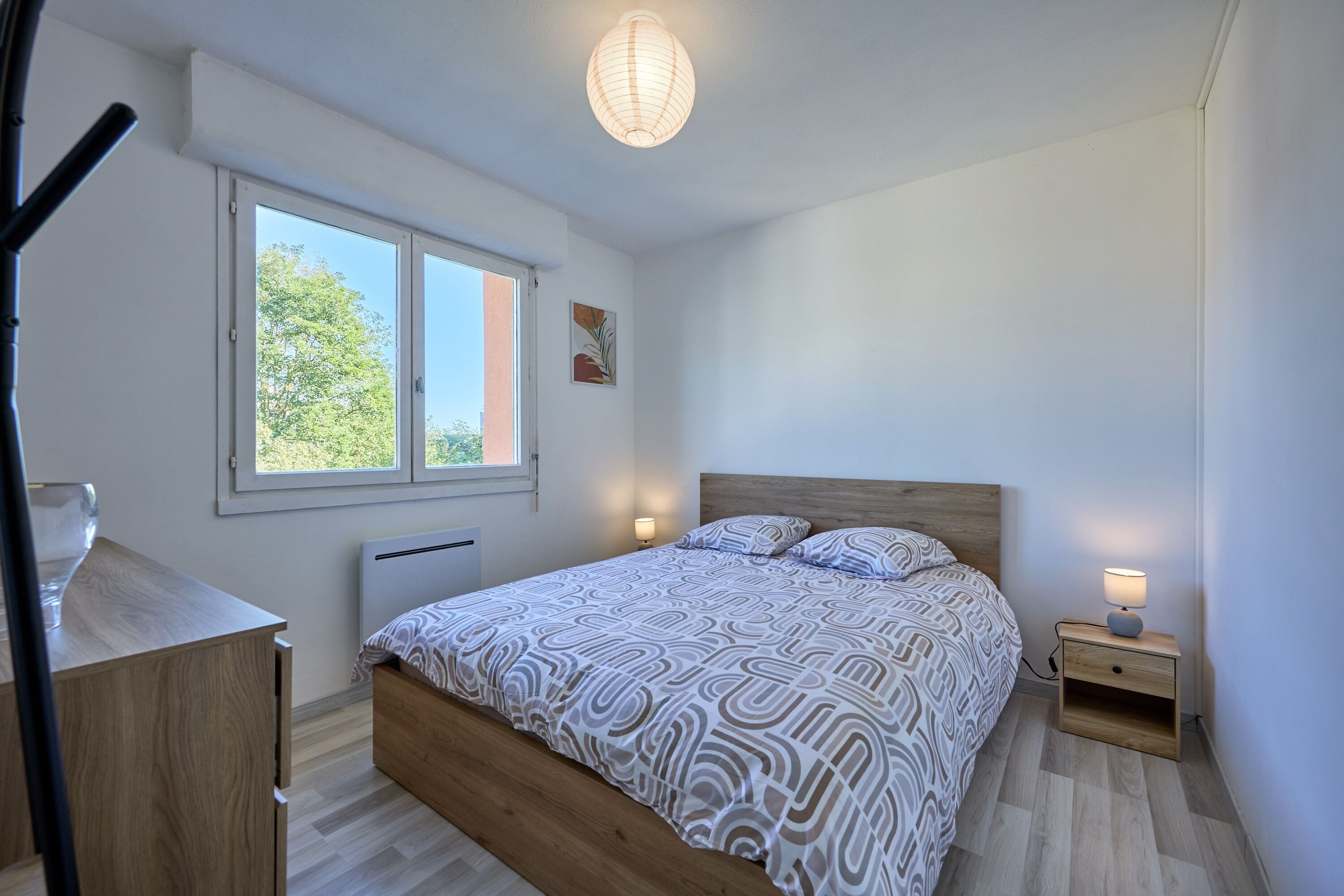 Ein Schlafzimmer mit einem hölzernen Bett, einem Nachttisch mit Lampe, einem Fenster mit Blick auf Bäume und einem Heizkörper unter dem Fenster.