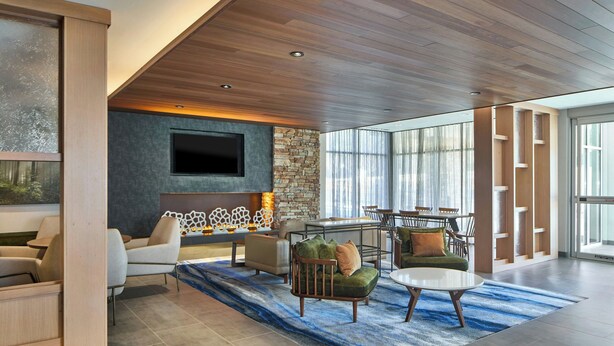 A modern living room with a stone wall, wooden ceiling, and a large TV. There are multiple seating areas with sofas and chairs, and a dining table. - Fairfield By Marriott Inn & Suites Cornelia (Cornelia)