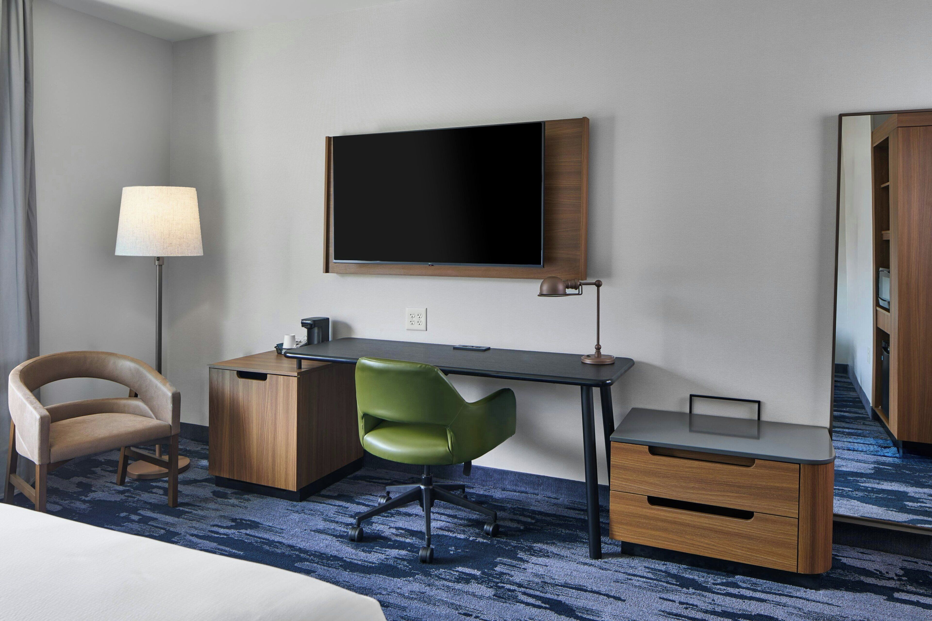 A hotel room with a bed, a desk with a chair, a TV, a lamp, and a mirror.