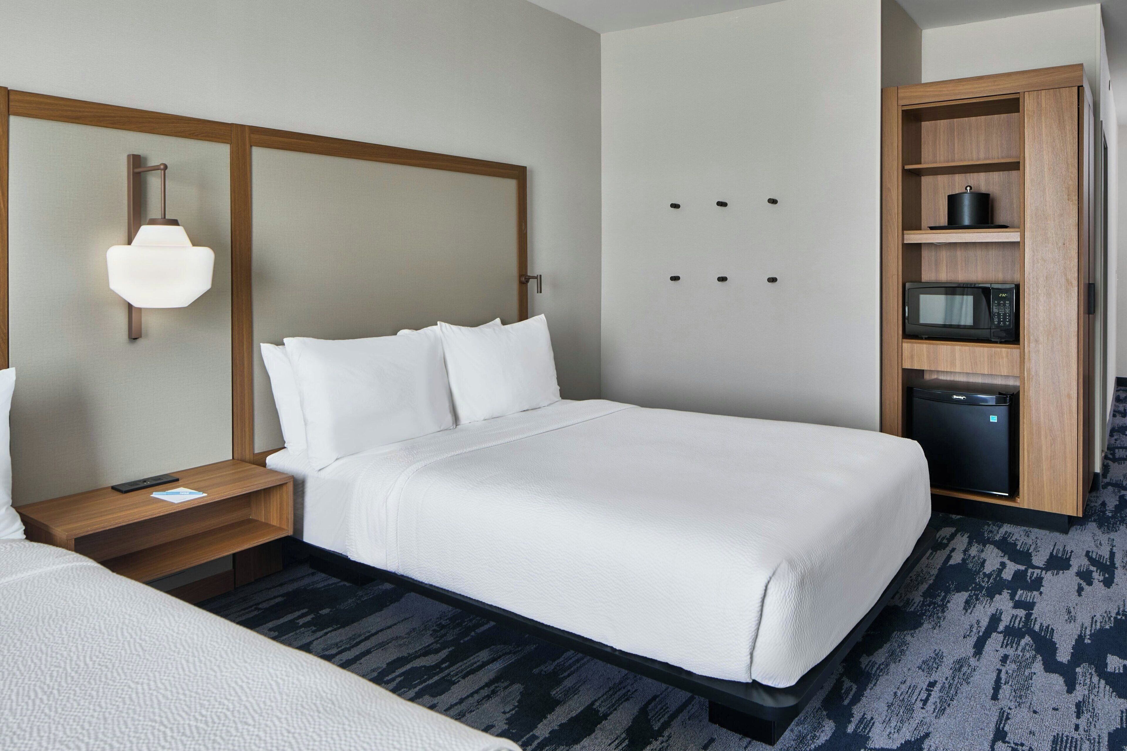 A hotel room with a large bed, a bedside table with a lamp, a built-in microwave, and a small refrigerator.