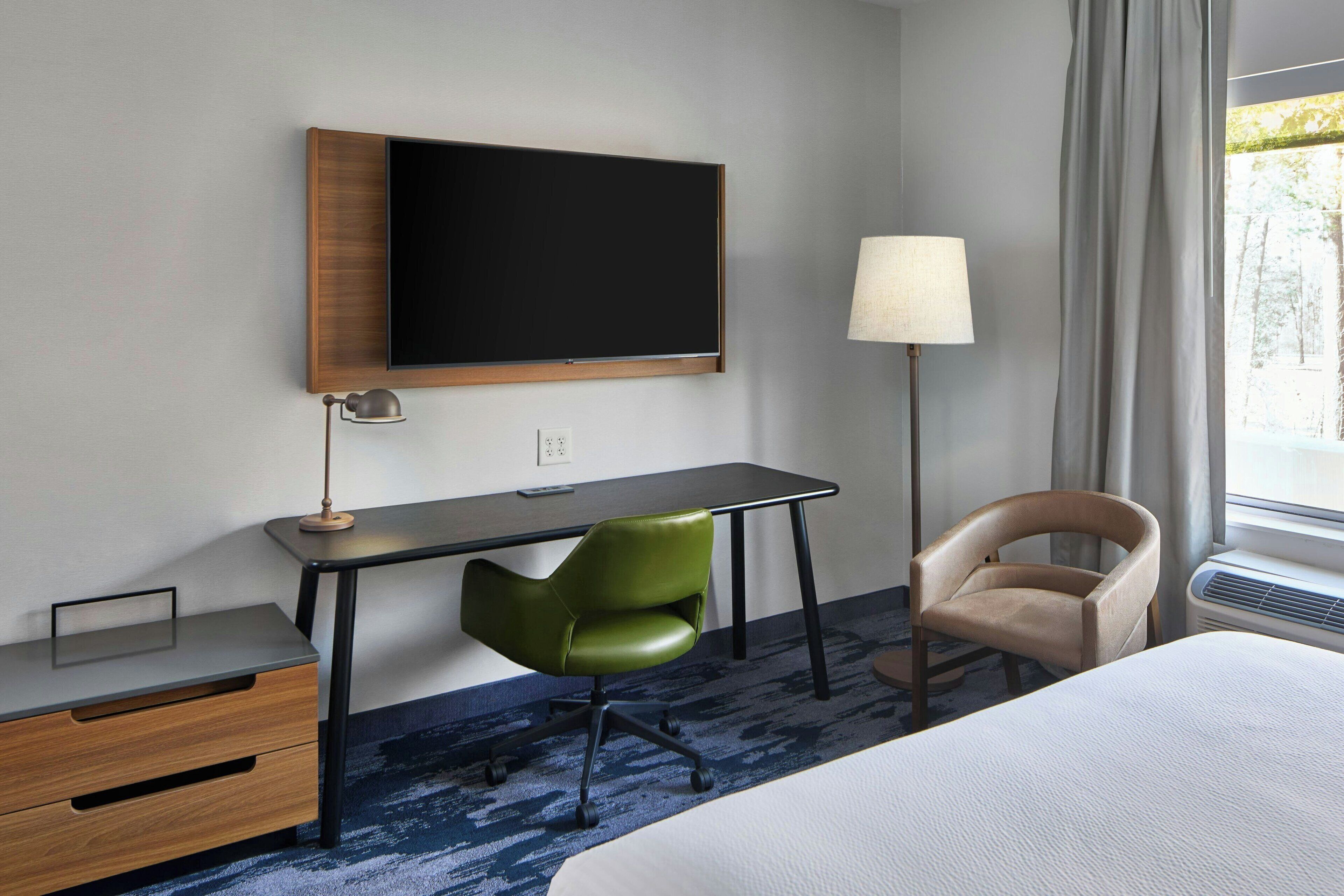 A hotel room with a bed, a desk with a chair, a television, a lamp, and a window with curtains.