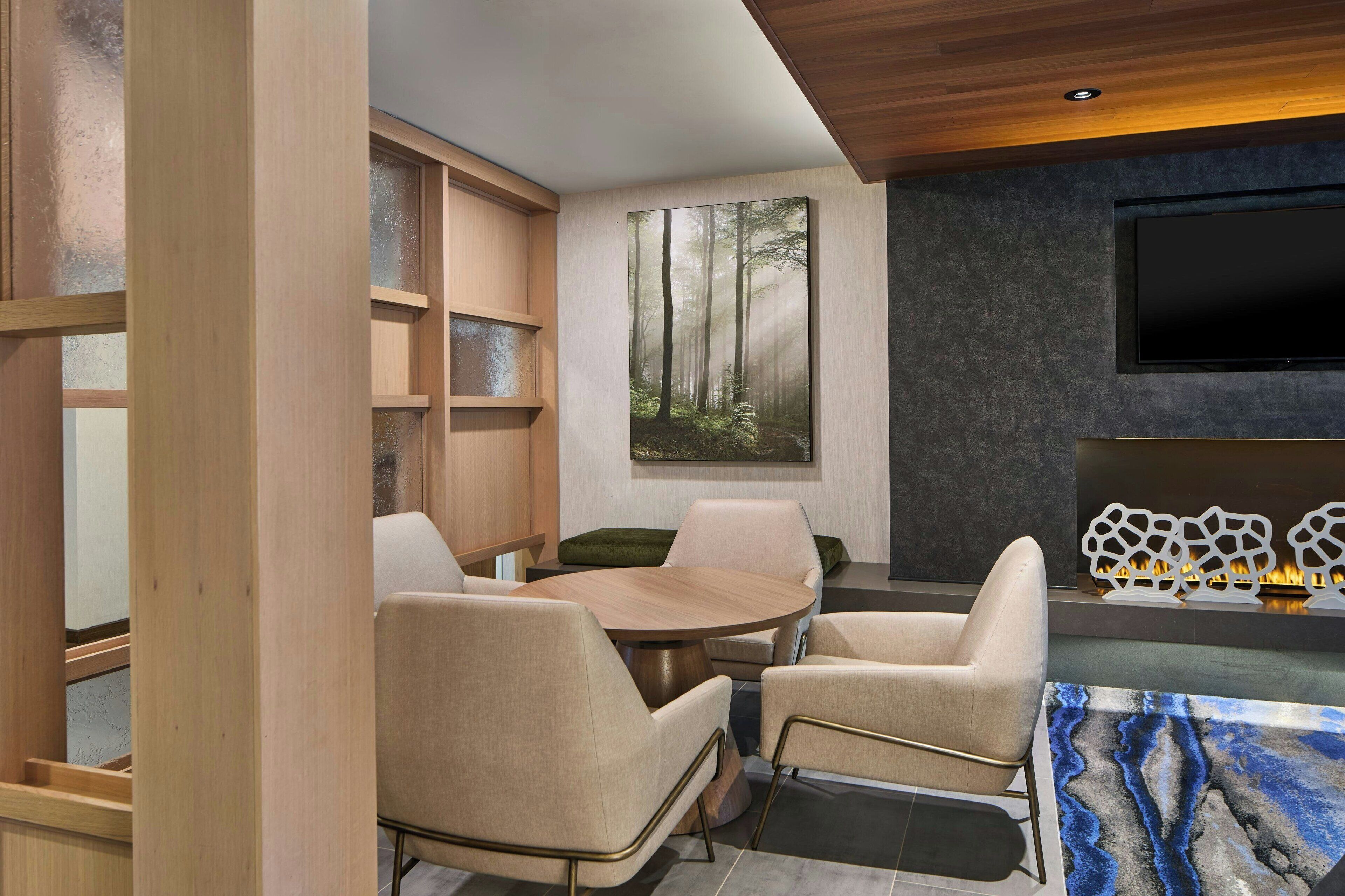 Una sala de estar moderna con una mesa redonda de madera, sillones color beige, un cuadro grande con temática de bosque, un televisor de pantalla plana y una chimenea decorativa.
