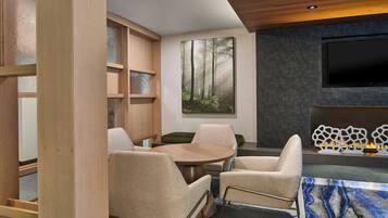 Una sala de estar moderna con una mesa redonda de madera, sillones color beige, un cuadro grande con temática de bosque, un televisor de pantalla plana y una chimenea decorativa.