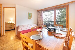 A dining area with a wooden table set for four, a sofa with a floral cushion, and a window with curtains. - Roche et Neige - Proche remontées mécaniques (Chamrousse)