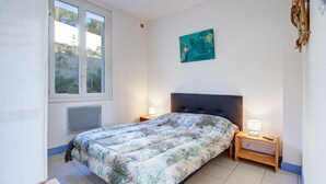 A bedroom with a bed, a nightstand, a wall-mounted lamp, and a window with a view of greenery.