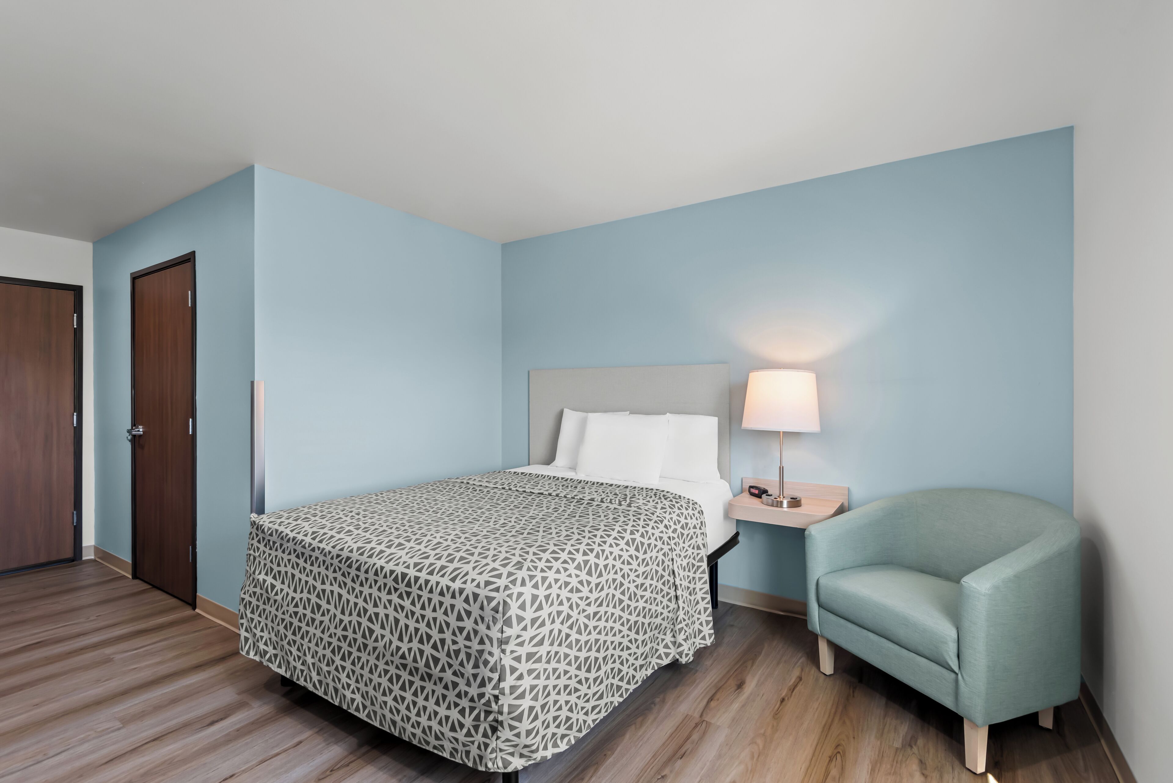 A hotel room with a bed, a nightstand with a lamp, a chair, and a door.