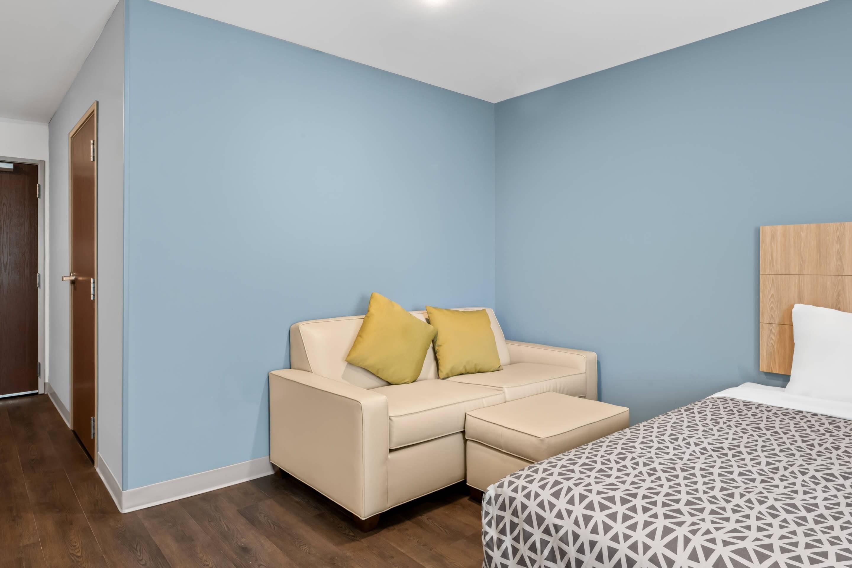 A hotel room with a beige sofa, a bed with a geometric-patterned bedspread, and a wooden headboard.