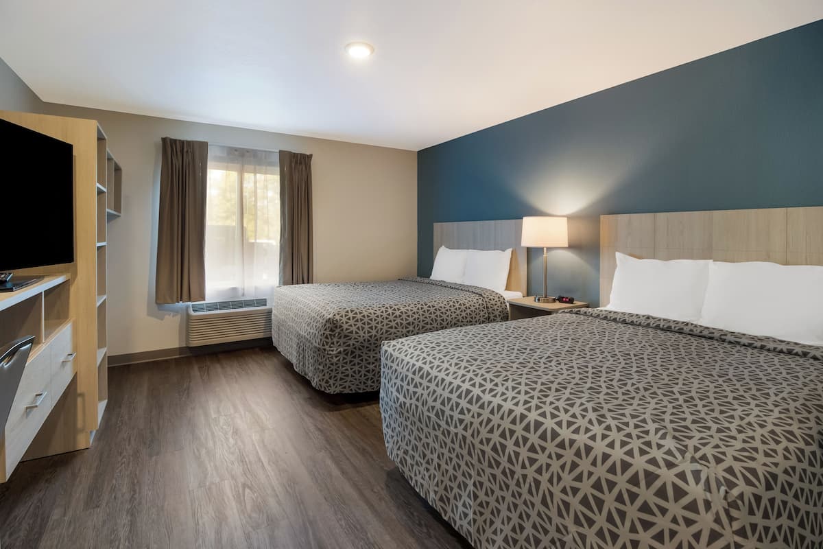 Une chambre d’hôtel avec deux lits, une télévision, un bureau avec des tiroirs, une fenêtre avec des rideaux et une lampe sur la table de chevet.