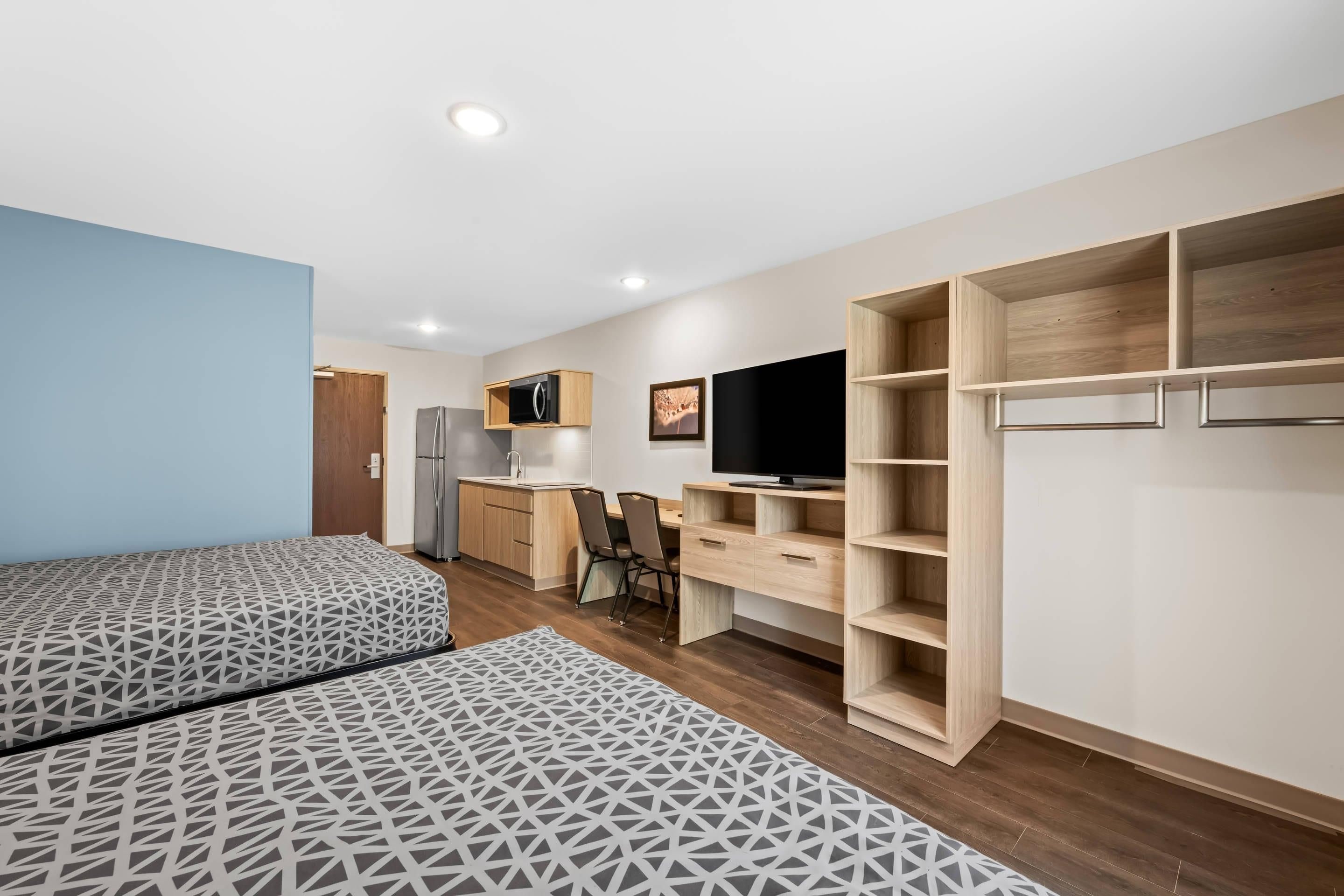 Una habitación funcional con dos camas, kitchenette, escritorio con silla y televisor.