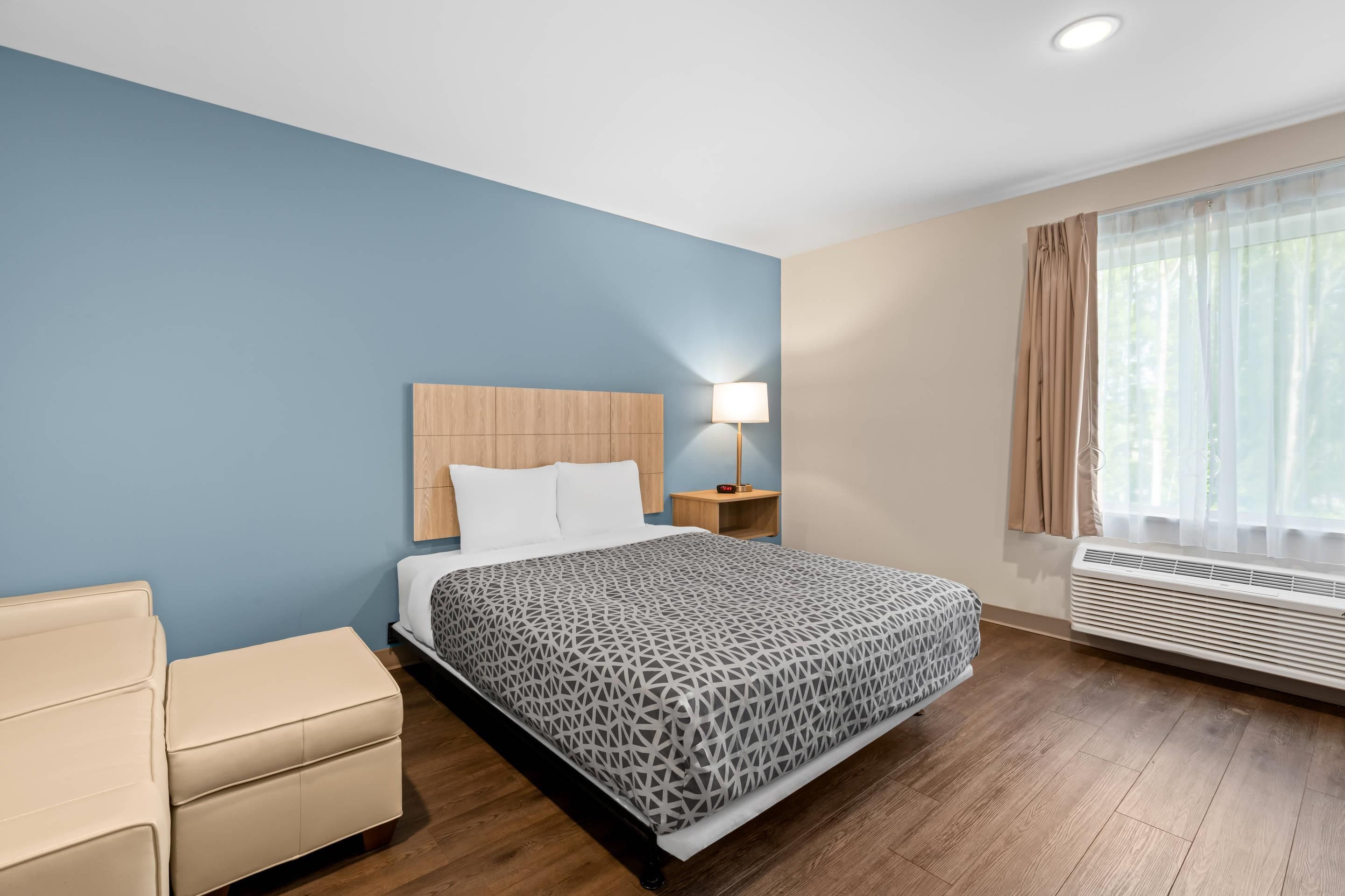 A hotel room with a bed, bedside table, lamp, air conditioning unit, and a chair.