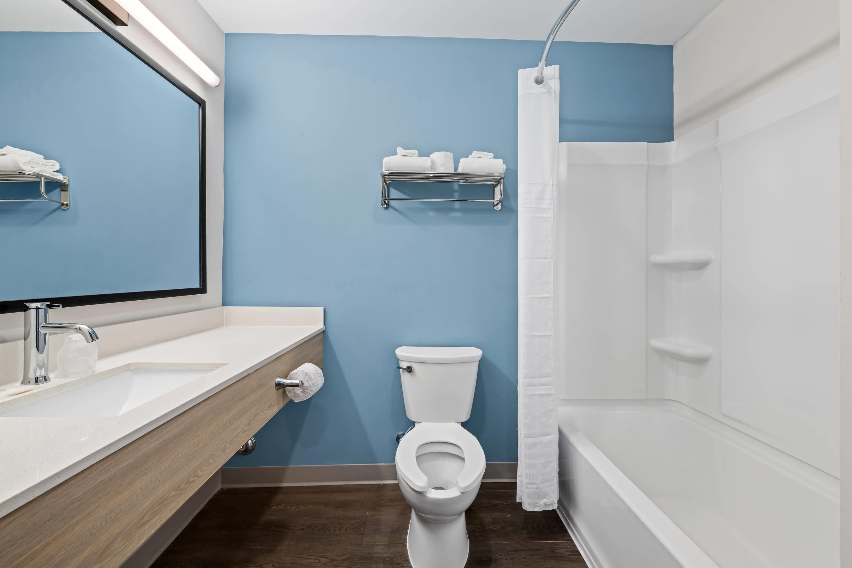 Une salle de bain avec des toilettes blanches, un lavabo avec un miroir, un rideau de douche et une étagère avec des serviettes.