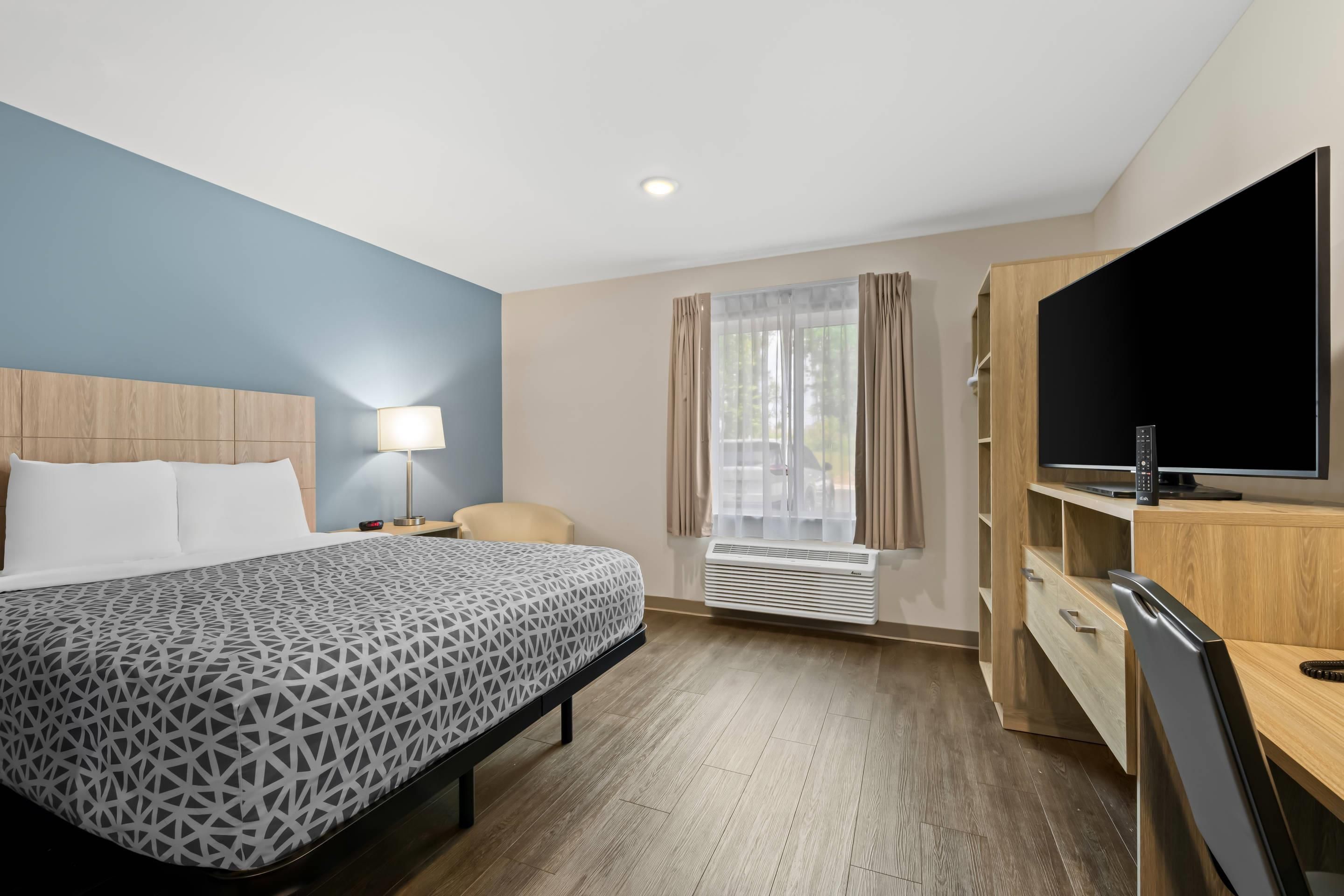 A hotel room with a bed, a desk with a flat-screen TV, a chair, a lamp, and a window with curtains.
