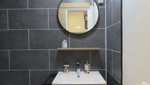 Un baño con paredes de baldosas oscuras, un espejo redondo, un lavabo blanco y dos tazas estampadas.