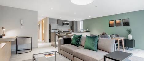 Una sala moderna con un sofá, una mesa de centro de vidrio y una mesita auxiliar. En el fondo, hay una zona de cocina con un microondas y una mesa de comedor.