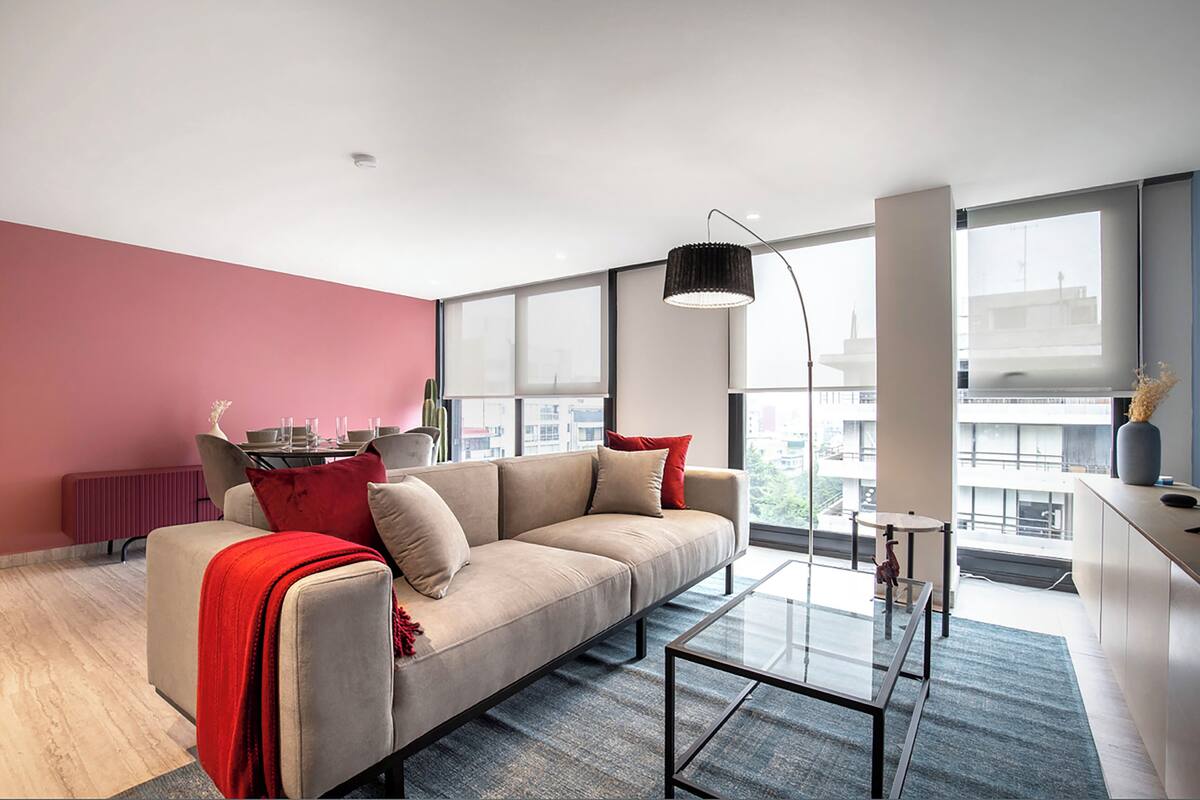 Ein modernes Wohnzimmer mit einem beigefarbenen Sofa, roten Kissen, einem Glastisch und einem großen Fenster mit Blick auf die Stadt.