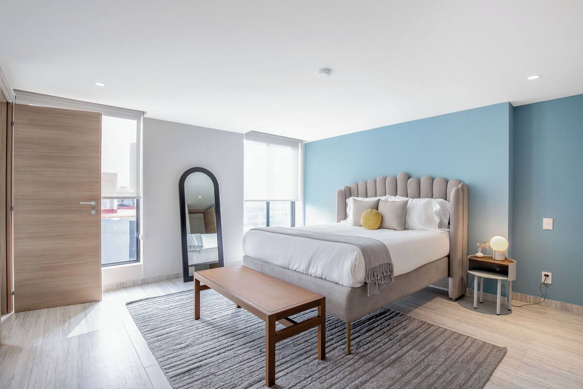 Ein modernes Schlafzimmer mit einem großen Bett, einem Nachttisch, einer Bank, einem Spiegel und einem Fenster mit Blick.