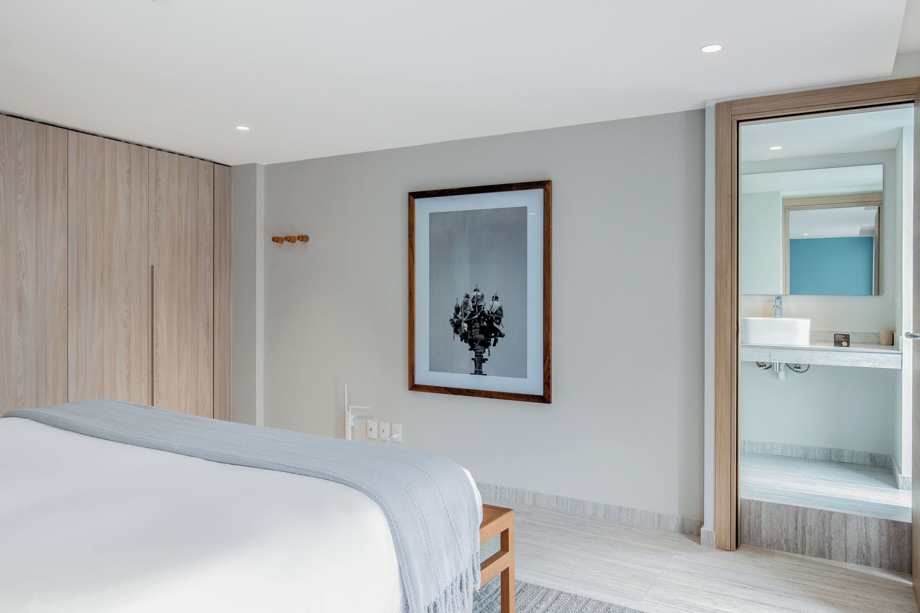 Ett modernt hotellrum med en stor säng, ett inramat konstverk på väggen och ett badrum med spegel och handfat.