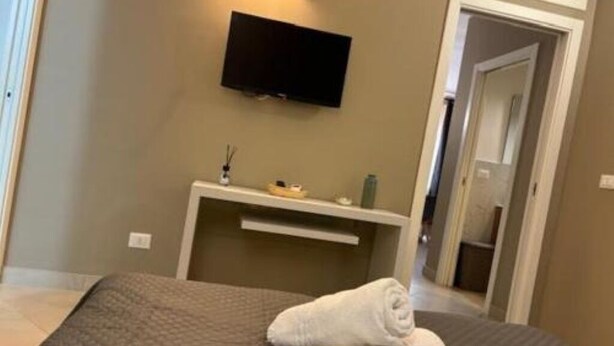 A hotel room with a bed, a flat-screen TV mounted on the wall, a small desk with a lamp, and a door leading to another room.