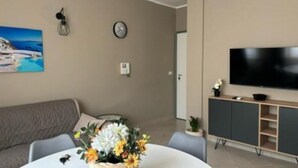 A modern living room with a grey sofa, a round dining table with a centerpiece, a flat-screen TV mounted on the wall, and a grey cabinet below.