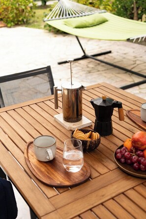 Un tavolo di legno con una caffettiera, una French press, una tazza, un bicchiere d'acqua, una ciotola di biscotti, un piatto di frutta e un'amaca verde sullo sfondo.