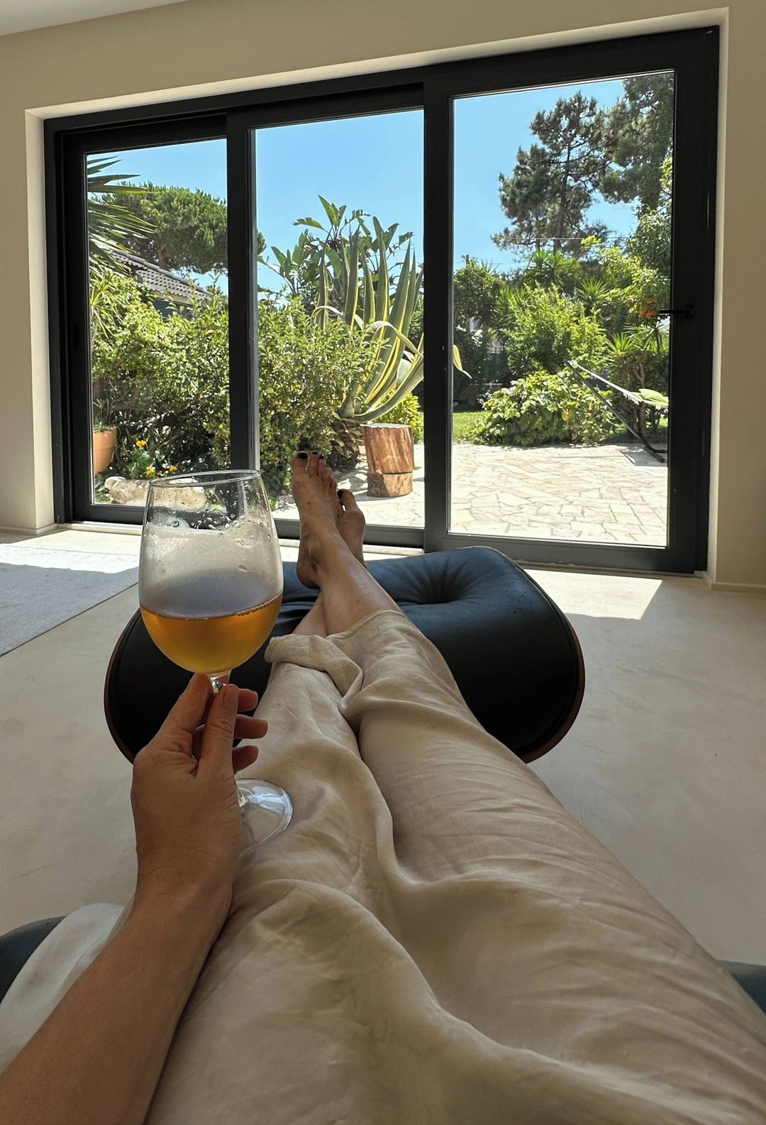 A person reclining on a black cushion with a glass of beer, feet propped up on a black ottoman, in a room with large glass doors leading to a garden with plants and a stone pathway.
