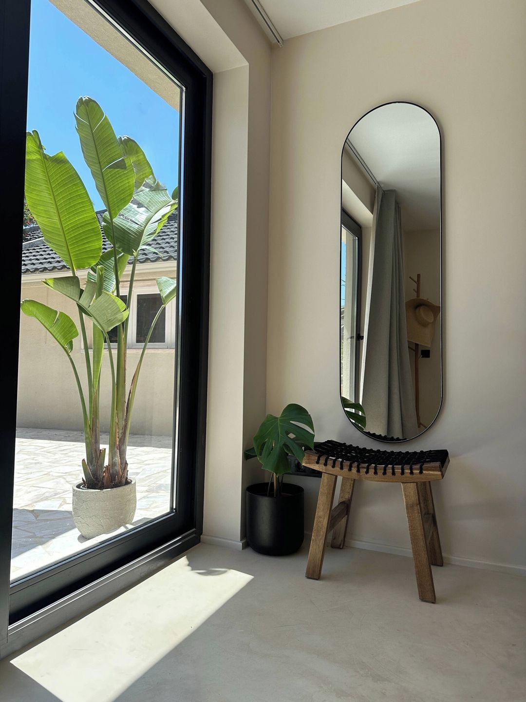 A room with a large window, a potted plant, a small wooden stool, and a mirror on the wall.