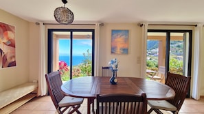 Ein Essbereich mit einem runden Holztisch, vier Stühlen, einer Blumenvase und Blick auf das Meer durch große Schiebetüren.
