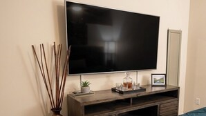 A bedroom with a large flat-screen TV mounted on a wooden console, a bed with a teal cover, and a vase with sticks beside the TV.