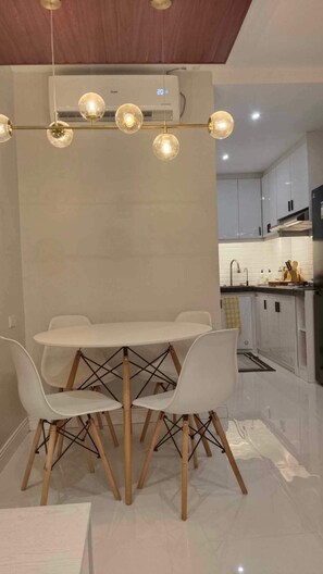 A modern dining area with a round table and white chairs, a ceiling-mounted light fixture, and a kitchen in the background.