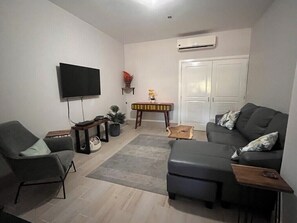 Ein Wohnzimmer mit Sofa, Sessel, Flachbildfernseher, kleinem Tisch und einer Tür.