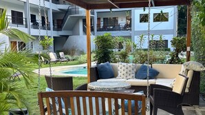 A covered outdoor seating area with wicker furniture, a small round table, and a view of a pool and building in the background. - Relax in Costa Rica Pura Vida Style and enjoy the magic! (Coco)